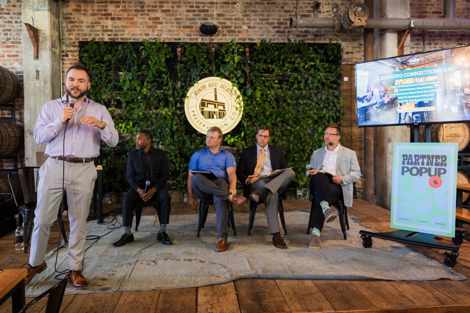 A speaker introducing a panel on the Monday Night Brewing stage in front of the green wall.